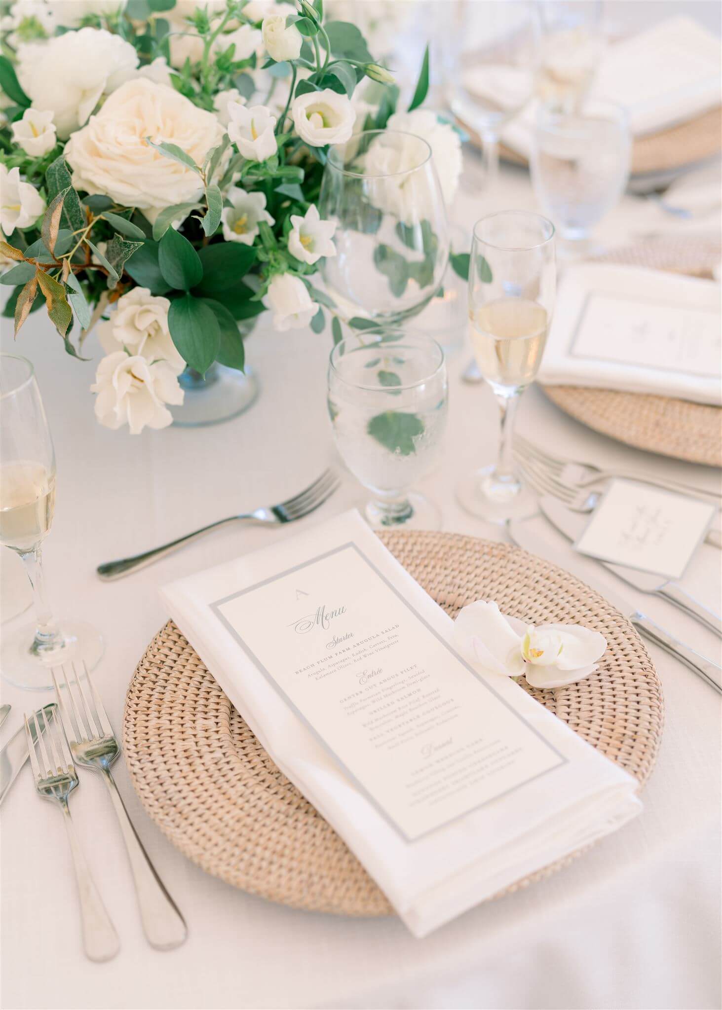Elegant white and blue wedding menu cards with script font and monogram sitting upon a woven rattan charger